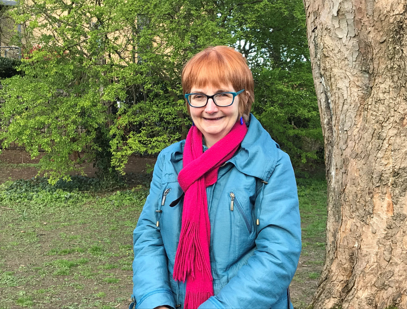 Sheila outside in a raincoat beside a tree, smiling