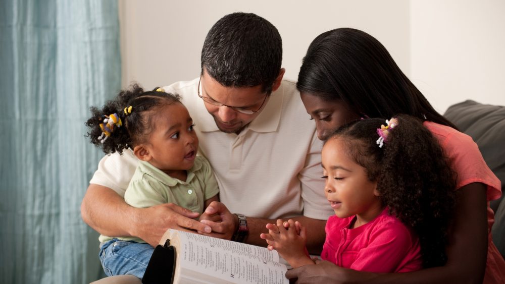 A family of 4, two parents and their young children all sat on the sofa together reading a book