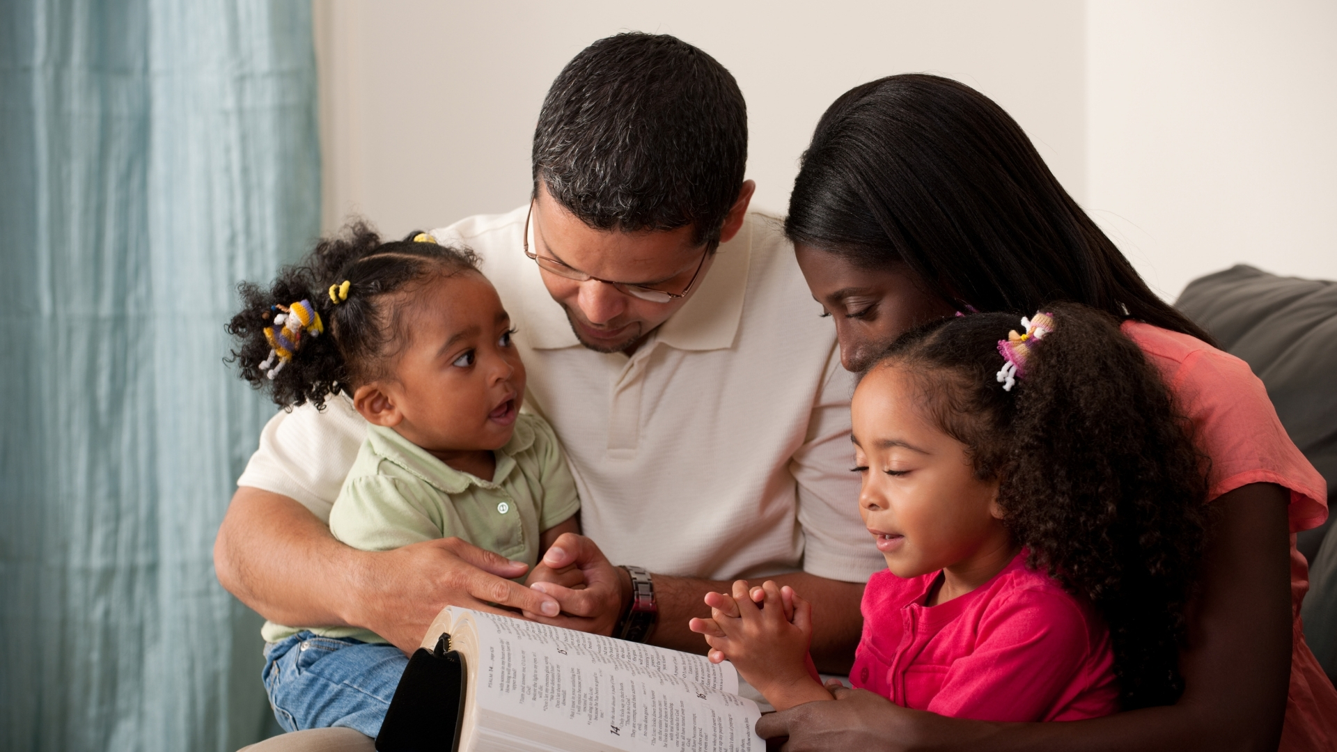 A family of 4, two parents and their young children all sat on the sofa together reading a book