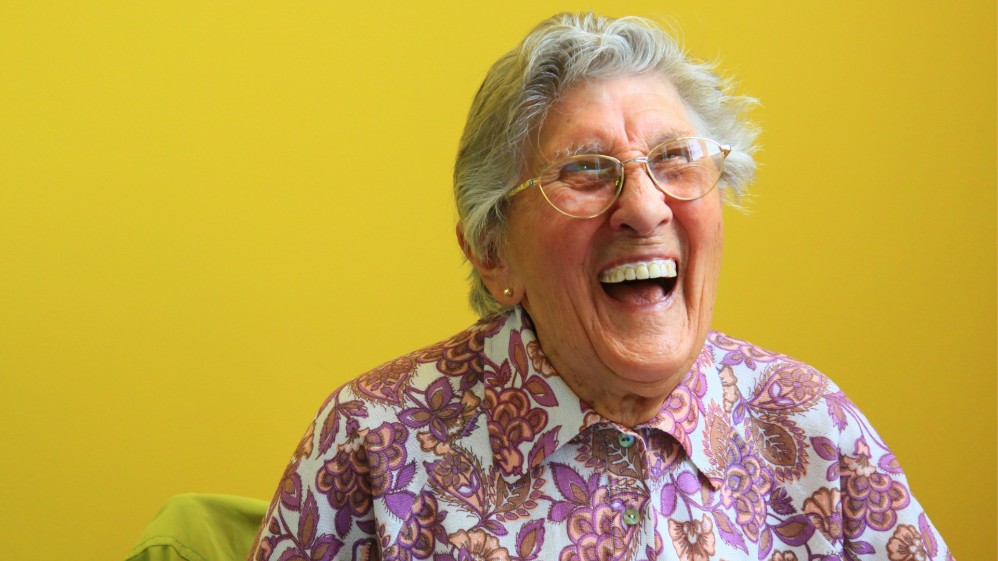 An elderly woman wearing glasses and a pink floral top laughs joyfully.