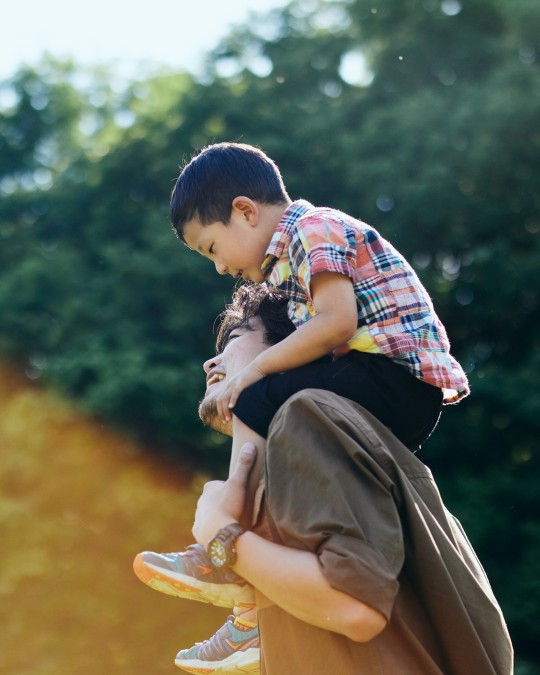 A man in a brown shirt smiles warmly as he looks up at a young boy riding on his back. The boy, wearing a checked shirt and black shorts, gazes down at the man with a joyful expression.