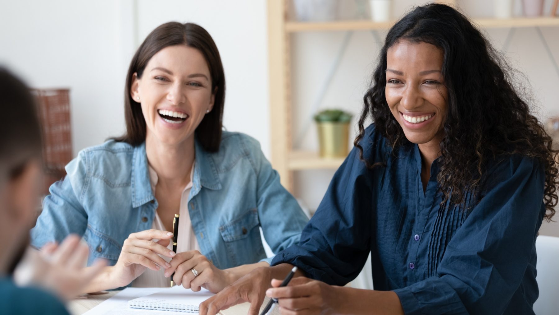 Two women seated at a table with pens and notepads, both smiling. One woman is white with shoulder-length dark hair, wearing a light blue denim shirt over a white top. The other woman is Black with long curly hair, dressed in a dark blue blouse. A third person’s hand is visible in the foreground, slightly blurred.