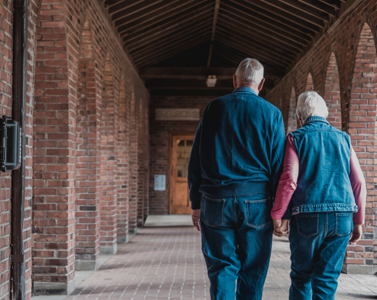 Elderly couple wearing denim walking down hallway holding hands