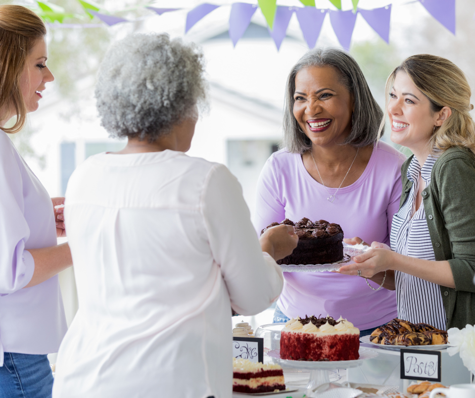 A group of people gathered around a table at what appears to be a casual celebration or party. The table is decorated with several cakes and pastries, including a chocolate cake being served and a red velvet cake with cream frosting. There are small signs on the table, and purple triangular bunting is hanging above as decoration. The setting looks bright and festive, likely outdoors or in a well-lit space.
